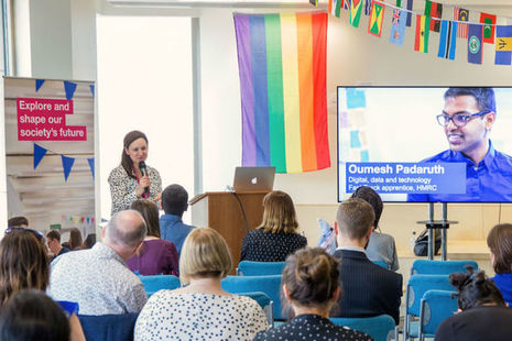Proffesional woman stood at a lectern talking with a microphone to a room full of people