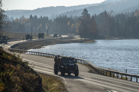 A Foxhound patrol vehicle leads a convoy of British Army trucks along winding road by the side of a Norwegian lake