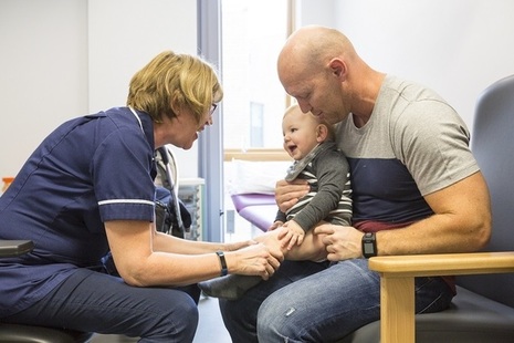 Baby sits on Father's knee while in appointment with nurse