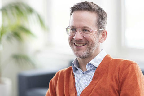 Head and shoulders of bespectacled man wearing a blue and white speckled shirt and orange cardigan