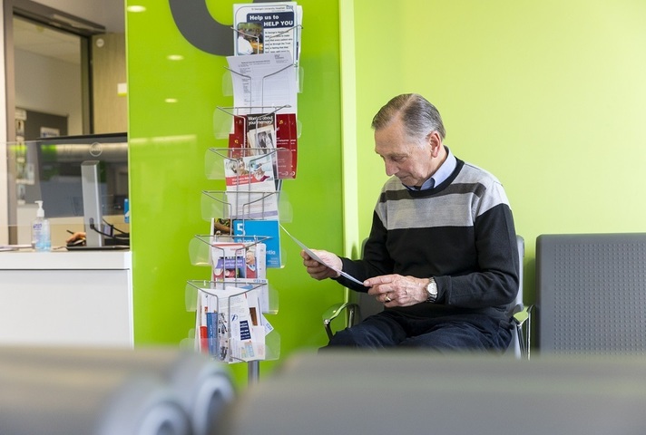 An older man sitting in a GP waiting area reading a leaflet