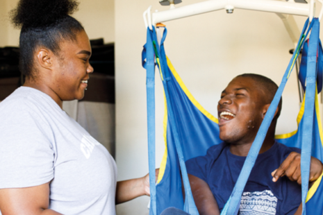 A social care worker laughs with her client who is sitting in a hoist.