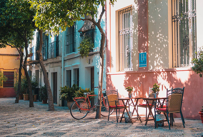 French street scene with colourful buildings, trees, table and chairs and a bicycle.