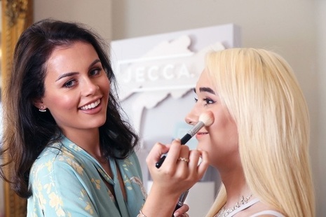 A photo of a lady smiling at the camera whilst doing another lady's makeup.