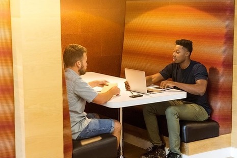 Two men sat opposite one another in an orange seating area, with a laptop computer between them.