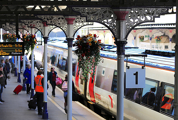 Rail station platform in summer, with passengers, a train and hanging baskets with flowers.