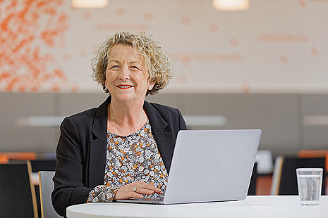 A lady sat at a desk behind and laptop smiling at the camera.