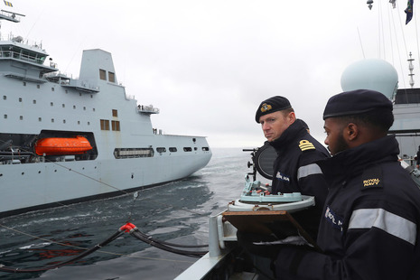 Image depicts two Navy personnel on a ship with the background of another ship.