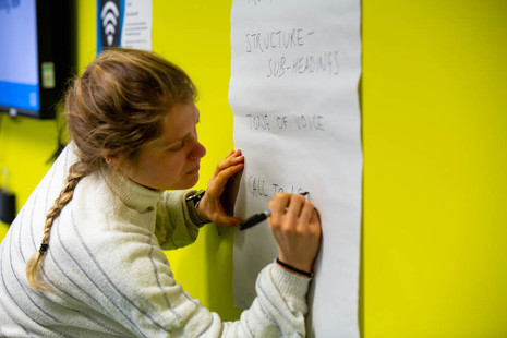 A woman writes 'tone of voice', 'structure - sub headings' and 'call to action' on a board during a content planning session.