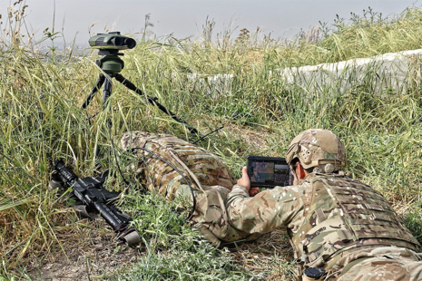 Soldier using the system, laying flat on the ground in long grass, looking into a tablet screen. 