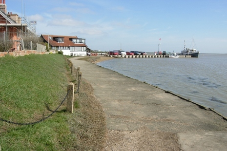 Image shows path curving from right next to the harbour past some houses towards a jetty with a boat moored at the end.