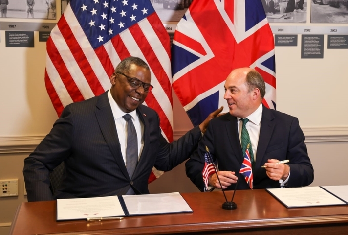Ben Wallce and Lloyd Austin smile as they sign the agreement extension in front of the UK and US flags