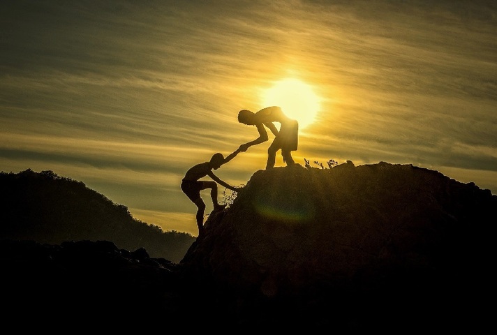 Silhouette of person helping companion climb on to a hilltop.