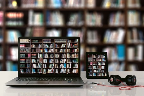 Image of laptop and mobile phone on desk both showing same picture of bookcase, which is also in the background.