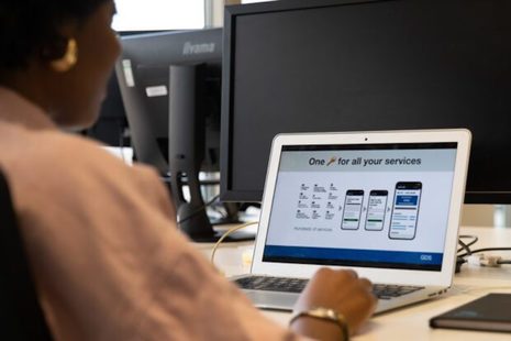 A person sits at a laptop. A presentation on screen shows a slide titled "One key for all your services" that shows government department logos and a user journey.