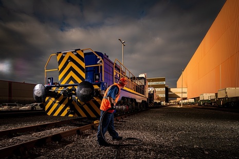Member of Sellafield's rail team holding large charging connection standing next to new hybrid locomotive on the Sellafield sites. There are industrial buildings in the background.