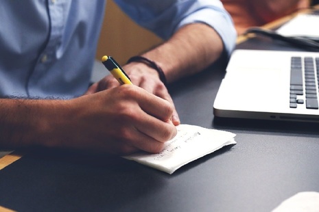 Man sitting at desk and writing on notepad next to laptop
