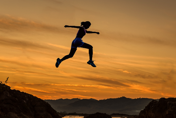 Female athlete silhouetted against sunset sky as she leaps across mountain ravine.
