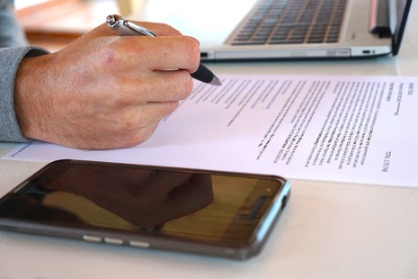 Image of hand holding pen above document on desk with laptop and mobile phone alongside