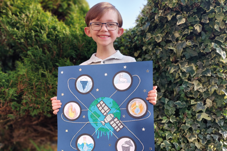 Competition winner Callum stands holding his winning design, a drawing of a satellite orbiting Earth with icons of climate change