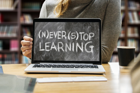 Image of woman sitting behind laptop screen which shows blackboard with chalk text which reads (N)ever (S)top Learning