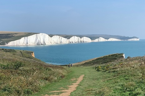 Image shows wide grass path sloping down towards a bright-blue sea, beyond which a line of white chalk cliffs can be seen stretching into the distance.