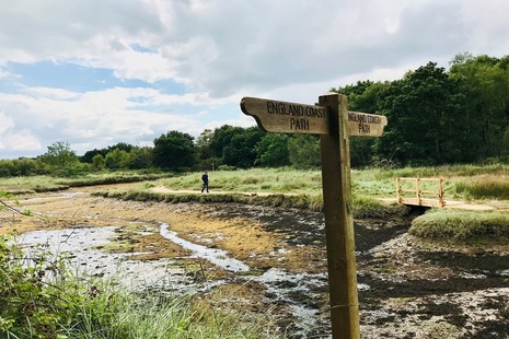 Image shows a person walking from left to right through countryside following a path beside a dried-up stream towards a small footbridge. The foreground is dominated by a sign pointing left and right to 'England Coast Path.