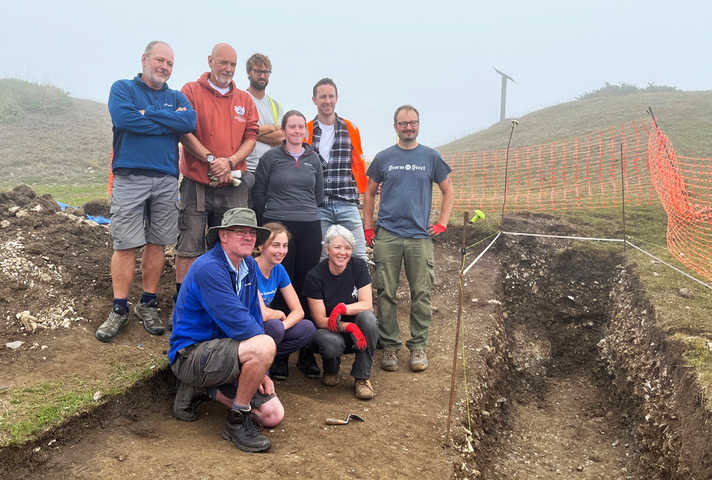 A group of men and women taking part of in an archaeological dig pose next to trenches dug on a hillside location on the Dorset coastline.