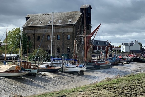 Photograph showing a line of sailing boats and barges resting on a river bed when the tide is out in front of an old warehouse.