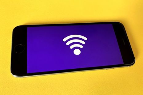 Мобильный телефон лежит горизонтально на желтом фоне.  Экран мобильного телефона имеет фиолетовый фон, а на фиолетовом фоне находится большой символ беспроводной связи.