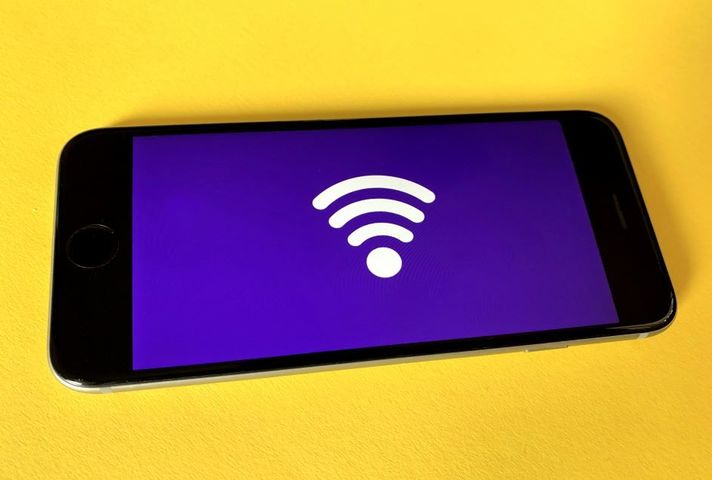 Mobile phone lieing horizontally against a yellow backdrop. The screen of the mobile phone has a purple backdrop and a big wireless reception symbol sits on the purple backdrop. 