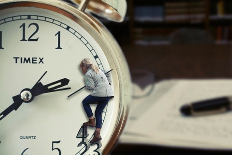 Woman climbing on clock face with diary on desk in background.