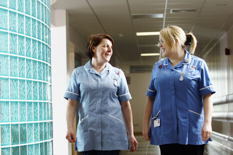 Two nurses wearing blue uniforms chatting while walking through a corridor