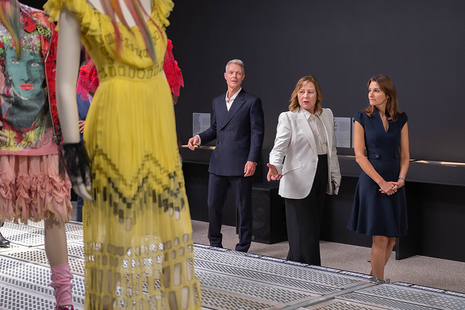 NewGen fashion designs including a yellow dress and a pink textured skirt are in the foreground, with Culture Secretary Lucy Frazer being shown round the display in the background