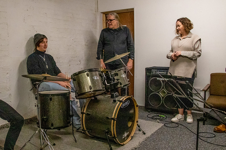 a teen plays a drumkit in a music room with two adults 