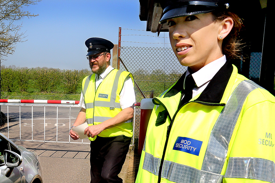 A male and female MOD guard in high visibility jackets stand outside a site entrance..