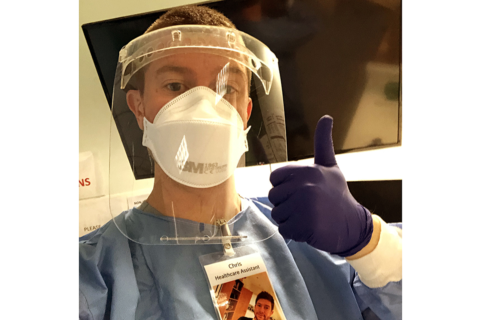 Midshipman Christopher Storer gives a thumbs up in his protective mask and apron. He wears a badge on his chest showing a picture of his face.