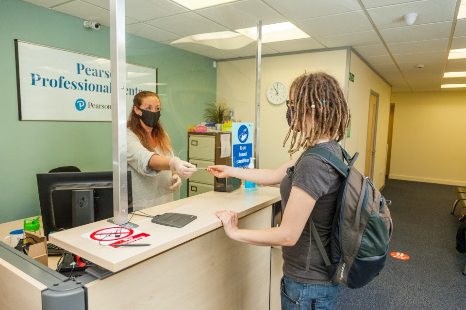 Photo of a woman handing her driving licence to a member of staff behind a desk. They are both wearing face coverings.