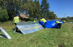 Workers wearing protective clothing assemble long metal barriers, which they cover in plastic sheeting, in a field with trees and a house to the left