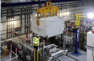 The image shows a white transport flask being moved within a building by an overhead crane.