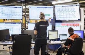 A woman standing up with her back to the camera looking at a selection of computer screens and talking to a male recruit also looking at the computer screens.