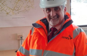 Mark standing in an florescent orange jacket and white hard hat with Amey written on the front smiling straight at the camera.