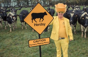 Herdsy - hero picture - Richard Hobson in a field with cows wearing a yellow suit and Herdsy's sign saying Caution - Farmers Making Money