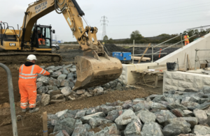 Image shows a digger to the left of the picture moving large rocks next to a concrete structure, overseen by a worker