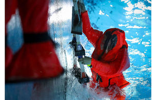 A sailor in a red diving suit, waist deep in water climbing up a wall using a rope.