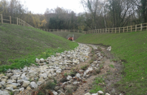 Image shows a dry stream bed curving from the left between green embankments towards a culvert