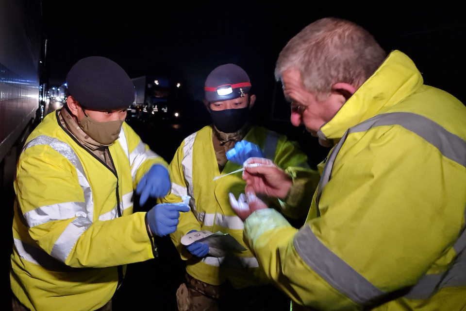 Three men standing around in the dark in high vis jackets holding COVID testing kits. 