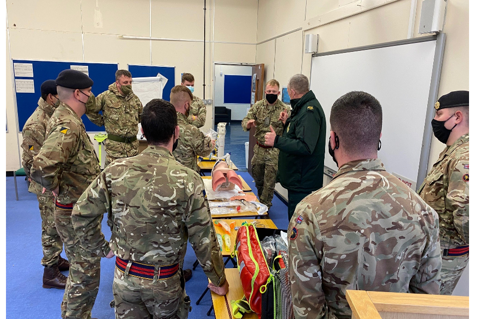 Members of the Armed Forces standing in a room receiving first aid training, listening to a man at the front demonstrate.