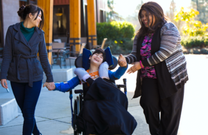 Two women hold the hands of a child using a wheelchair. Everyone is laughing and smiling.
