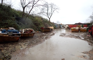 Image shows a muddy yard enclosed by a high bank on the left, full of skips packed with rubbish